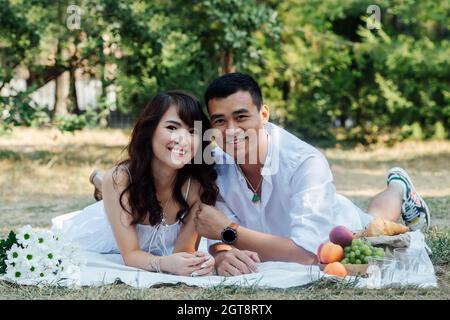 Un couple asiatique souriant se fait un pique-nique dans un parc, allongé sur leurs estomacs. Homme et femme en vêtements blancs dans une ombre d'arbre. Banque D'Images