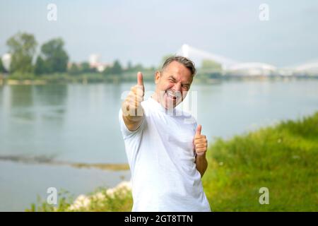 Un homme exubérant excitant donnant un geste triomphant avec un sourire heureux alors qu'il se tient sur la rive d'une rivière large sous le soleil du printemps Banque D'Images