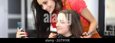 Jeune femme avec coiffeur assis au salon de beauté et prenant selfie sur smartphone Banque D'Images