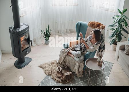 Une jeune femme dans un fauteuil confortable et un écossais assis près de la cheminée avec un chat domestique, boire du thé à la maison Banque D'Images