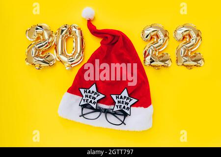 Ballons en aluminium sous la forme de numéros 2022 et chapeau de père Noël avec lunettes de fête sur fond jaune.Célébration du nouvel an. Banque D'Images