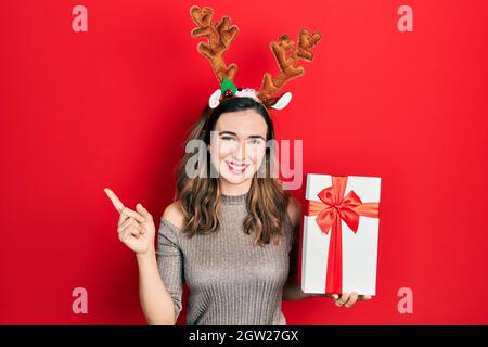 Jeune fille hispanique portant le chapeau de noël cerf tenant le cadeau souriant heureux pointant avec la main et le doigt sur le côté Banque D'Images