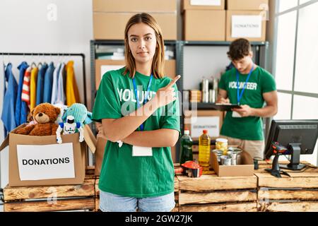 Jeune fille blonde portant le t-shirt de bénévole au stand de don pointant avec le doigt de la main sur le côté montrant la publicité, sérieux et calme visage Banque D'Images