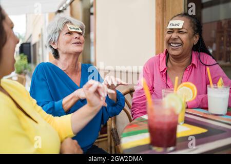 Amis séniors multiraciaux ayant du plaisir à jouer ensemble deviner qui jeu de front au bar restaurant - Focus sur le visage de femme africaine Banque D'Images