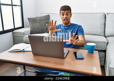 Jeune homme hispanique beau utilisant un ordinateur portable assis sur le sol peur et terrifié avec l'expression de peur stop geste avec les mains, criant dans le choc. pa Banque D'Images