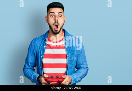 Jeune homme hispanique avec la barbe jouant le jeu vidéo tenant contrôleur peur et choqué avec la surprise et l'expression stupéfaite, la peur et le visage excité. Banque D'Images