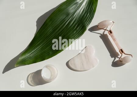 Grattoir de gouache rose, rouleau, pot de crème de feuille verte sur une table blanche. Équipement pour l'auto-massage et les soins de la peau pour le visage et le cou Banque D'Images