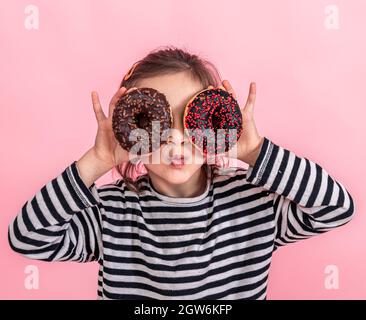 Portrait d'une petite fille souriante brunette avec et deux beignes appétissantes dans ses mains, ferme ses yeux avec des beignes, sur un fond rose. Banque D'Images