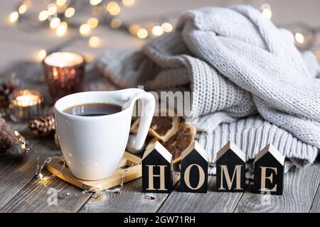 Composition hivernale confortable avec une tasse de thé, le mot décoratif Home, des éléments tricotés et des lumières bokeh. Banque D'Images