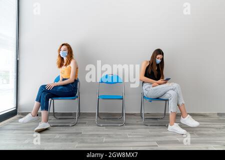 Deux jeunes femmes avec masque de protection sont assises dans une salle d'attente à distance. La rupture du virus Corona. Photo de haute qualité Banque D'Images