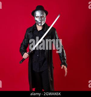 Un mystérieux assassin ninja dans un style noir. Un homme en vêtements noirs avec un cape et un chapeau, couvre son visage avec un masque et utilise un katana. Photo sur rouge Banque D'Images