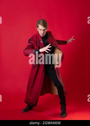 Un jeune homme scandaleux dans un manteau rouge audacieux dans un style noir vintage, pose épique et fait un geste de main. Photo sur fond rouge Banque D'Images