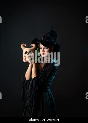 Une belle jeune sorcière tient un crâne de démon corné près de son visage et vous regarde avec un regard malicieux, une invitation à une fête d'Halloween, un costume de sorcière Banque D'Images