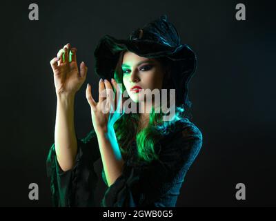 Une sorcière crée la magie, une jeune belle brune dans un chapeau pointu fait des passes avec ses mains, sorts. Portrait en gros plan, rétroéclairage vert Banque D'Images