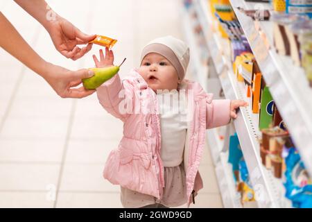 Shopping en famille. Un enfant mignon est debout près de l'étagère avec des produits, et choisira entre un bonbon et une poire, que la maman offre. Le concept de s. Banque D'Images