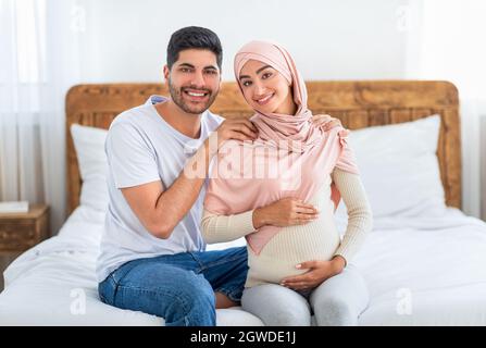 Heureux de s'attendre à ce que le couple arabe se câlin tout en passant du temps ensemble à la maison, de l'espace libre. Couple musulman enceinte assis au lit et souriant à la caméra Banque D'Images