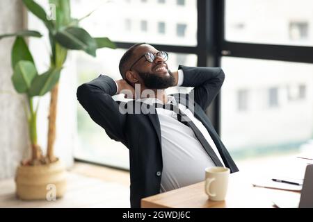 Portrait d'un homme d'affaires noir souriant et détendu en lunettes, penché en arrière sur la chaise, tenant les mains derrière la tête, se reposant au bureau moderne après avoir terminé le travail Banque D'Images