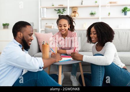 Une famille noire heureuse de trois personnes jouant avec un enfant, une maman et un papa attentionnés souriant et dessinant sur papier avec une petite fille en utilisant du penc coloré Banque D'Images