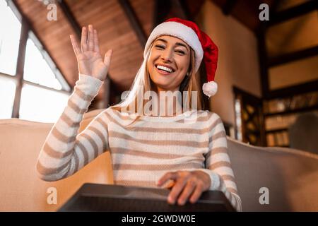 Concept d'accueil. Portrait d'une jeune femme excitée dans un chapeau du père noël en ordonnant bonjour avec la main regardant l'appareil photo, à l'aide d'un ordinateur portable à la maison. Une femme joyeuse dit Banque D'Images