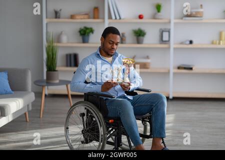 Un sportif noir paraplégique malheureux en fauteuil roulant tenant des trophées, se sentant déprimé à l'intérieur. Portrait d'un homme noir handicapé avec des prix exceptionnels Banque D'Images