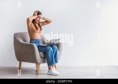 Concept d'échéance. Portrait d'une femme caucasienne choquée assise dans un fauteuil avec ordinateur portable et saisissant sa tête contre le mur blanc de studio, copie s. Banque D'Images