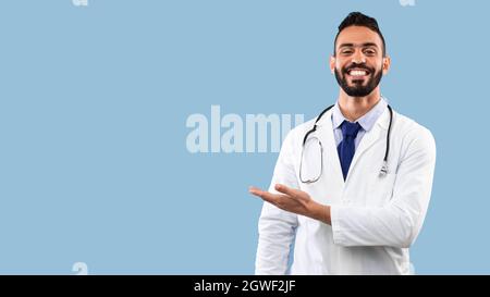 Happy Arabic Doctor Man souriant montrant l'espace de copie avec la main Publicité votre texte sur l'arrière-plan Blue Studio. Portrait du médecin ayant réussi. Medi Banque D'Images