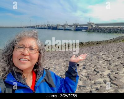 Souriante et mature touriste, Oosterscheldekering partie de Delta Works en arrière-plan, système de défense contre les inondations. Cheveux longs gris curly, blouson bleu, clo Banque D'Images