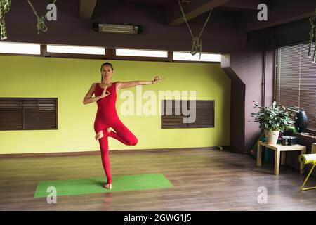 Une femme effectue un exercice vrikshasana avec un geste de conscience, pose d'arbre, se tient sur un tapis dans le studio, s'entraîne dans une combinaison rouge de sport Banque D'Images