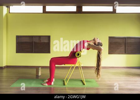 Une femme pratiquant la thérapie de yoga, exécute un coude arrière, mettant un tapis sous le bas du dos, les trains assis sur une chaise, dans le studio dans un rouge sports ju Banque D'Images