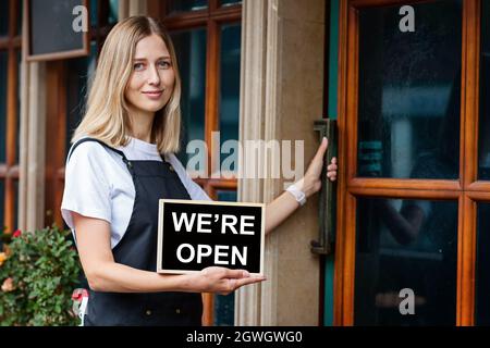 Jeune femme caucasienne de 30 ans avec des cheveux blond courts tenant une pancarte ouverte près des portes du café. Rouvrir le restaurant après le coronavirus Covid-19 Banque D'Images