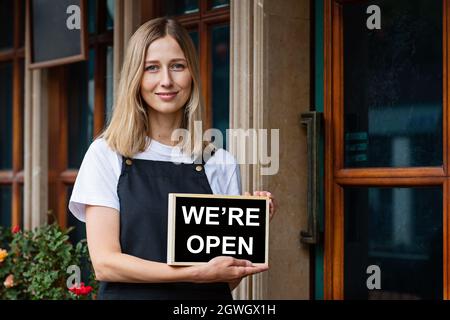 Jeune femme caucasienne de 30 ans avec des cheveux blond courts tenant une pancarte ouverte près des portes du café. Rouvrir le restaurant après le coronavirus Covid-19 Banque D'Images