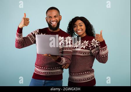 Noël shopping en ligne. Couple afro-américain heureux avec ordinateur portable montrant le pouce vers le haut, posant ensemble dans les mêmes chandails chauds sur le dos bleu Banque D'Images