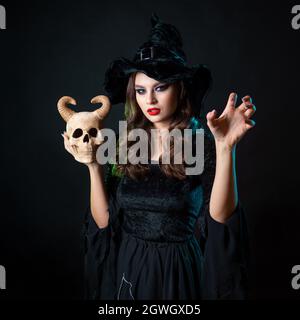 belle sorcière dans un chapeau pointu tient un crâne du démon, dans ses mains. Fête d'Halloween, Sabbat des sorcières. Jeune belle brunette en costum sorcière Banque D'Images