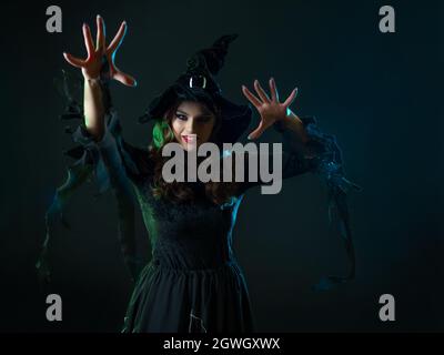 Une sorcière crée la magie, une jeune belle brune dans un chapeau pointu fait des passes avec ses mains, sorts. Fête du costume d'Halloween Banque D'Images