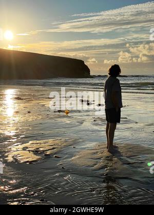 Vue arrière sur toute la longueur d'un homme mûr se tenant sur la plage en fin d'après-midi tandis que le soleil se couche. En regardant vers l'horizon, porter un short et un t-shirt. Banque D'Images