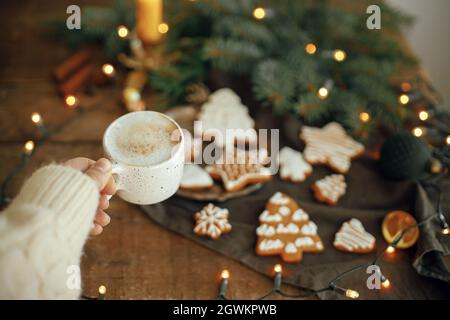 Main tenant une tasse de café chaud sur fond de biscuits de pain d'épice de noël, branches de sapin, lumières chaudes sur la serviette et table rustique en bois. Bonjour hiver Banque D'Images