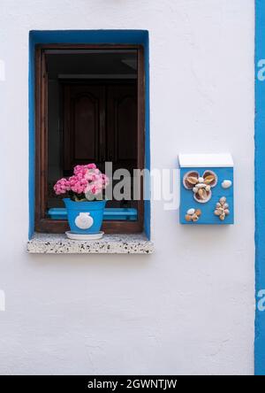 fenêtre bleue sur une façade blanche d'une maison dans un village de pêcheurs, avec un pot de fleurs violettes et une boîte aux lettres bleue avec ornements de seashell, verticale Banque D'Images