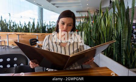 Gros plan sur une belle femme hispanique lisant le menu assis devant une table dans un restaurant orné de plantes Banque D'Images