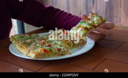 Main de femme prenant un morceau rectangulaire de pizza carrée avec du jambon, du poivron vert, de la tomate, des olives et du fromage d'une assiette blanche sur une table en bois Banque D'Images