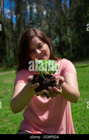 Belle jeune femme hispanique tenant une petite plante dans ses mains de champ avant d'être plantée dans un champ vert entouré d'arbres pendant la matinée Banque D'Images