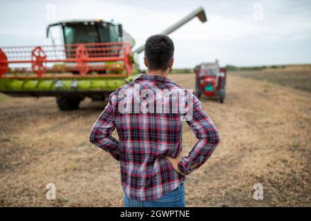 Vue arrière d'un jeune agriculteur attrayant se tenant dans un champ de soja récolté et regardant la moissonneuse-batteuse et le tracteur Banque D'Images
