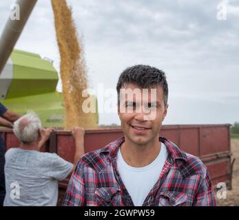 Jeune agronome attrayant debout dans le champ de récolte regardant la caméra. Deux agriculteurs expérimentés remplissent la remorque du tracteur avec des graines de soja provenant de la récolteuse. Banque D'Images
