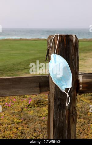 Le masque chirurgical bleu jeté est suspendu à un poteau de clôture abîmé sur un sentier de plage. Banque D'Images