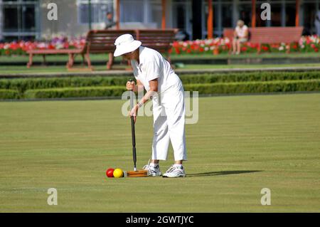 Une femme habillée de blancs appropriés utilise un maillet de croquet pour frapper des balles rouges et jaunes pendant la compétition dans les jardins gouvernementaux de Rotorua, en Nouvelle-Zélande, à l'été 2005. Banque D'Images