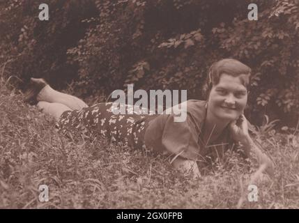 rarement vu la beauté sur la photographie des années 1930. Une jolie dame est couché sur l'herbe dans la nature. Elle sourit, elle est donc si heureuse, le temps est beau comme elle. Elle a une grande bouche, a une grosse mâchoire / jowl cheveux courts (très style Joséphine Baker) plus de grandes boucles d'oreilles en perles baroques, pendentif, gouttes d'oreille. Période: Années 1930 Source: Photographie originale Banque D'Images