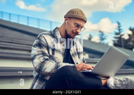 Homme sérieux barbu en lunettes travaille sur pc, indépendant assis dans le parc sur les escaliers, directeur de l'éducation Banque D'Images