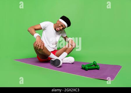 Photo pleine longueur de fatigué bouleversé peau sombre gars habillé sport ensemble assis tapis jambe stretching isolé couleur verte arrière-plan Banque D'Images