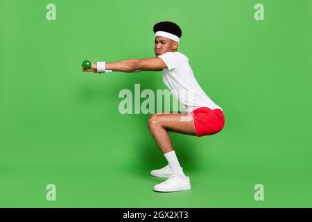 Photo pleine longueur de sérieux fatigué peau sombre homme porter t-shirt blanc squating avec des haltères isolé couleur verte arrière-plan Banque D'Images