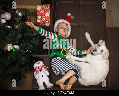 Un enfant en costume d'orf et un chapeau de père noël, se trouve à côté d'un chien blanc sur le canapé près de l'arbre de Noël. Joyeux Noël positif confortable atmos Banque D'Images
