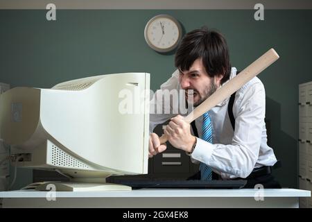 Un homme en colère détruit un vieux ordinateur avec une batte de baseball au bureau. Banque D'Images